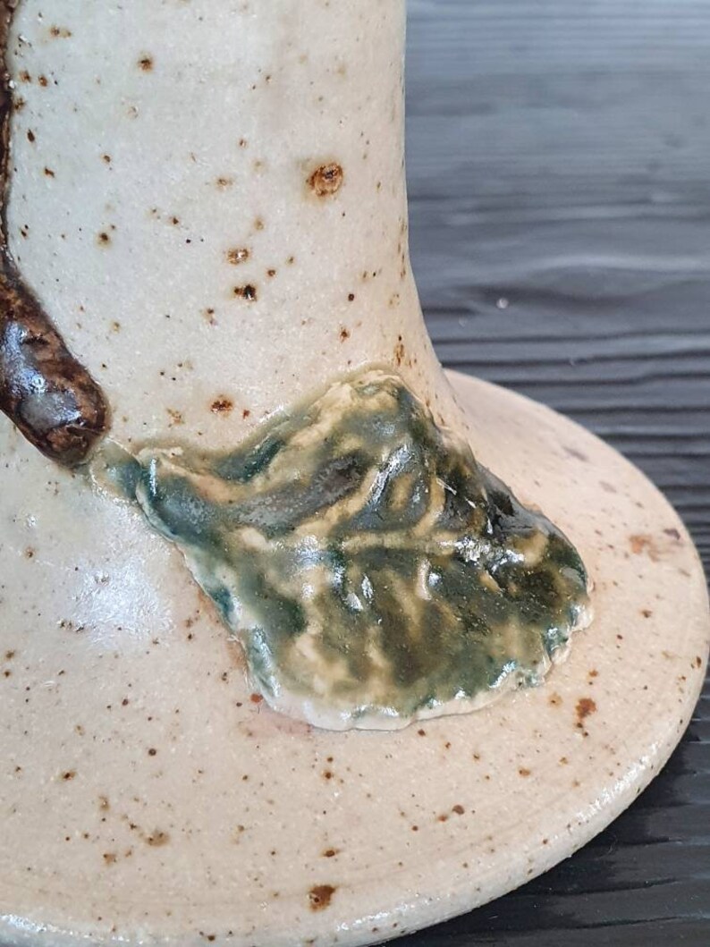 May include: A ceramic vase with a speckled white glaze and a green leaf design. The vase has a wide base and a narrow neck.