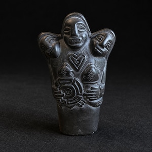 Peut inclure: Une sculpture en pierre gris foncé présentant trois visages sculptés au sommet, avec une forme de cœur centrale et un emblème circulaire tenu par des mains au milieu. La surface présente des lignes gravées et des motifs géométriques.