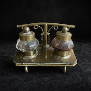 Pode incluir: Um conjunto de tinteiro de secretária em latão com uma base retangular e quatro pés pequenos, suportando dois tinteiros de vidro com tampas de latão. Um arco central em latão com volutas decorativas e pequenas correntes une os dois recipientes.