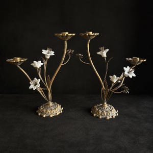 May include: A pair of gold-toned metal candelabras featuring a floral design with white ceramic lily-shaped accents. Each candelabra has a textured, leaf-patterned circular base and multiple stems that support candle holders at varying heights.