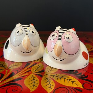 以下が含まれることがあります： 2つのセラミック製のフクロウ型ソルト＆ペッパーシェーカー。1つのフクロウは白と黒の模様、もう1つはピンクと黒の模様です。どちらもベージュのくちばしと笑顔の顔をしています。シェーカーは赤い表面にあります。