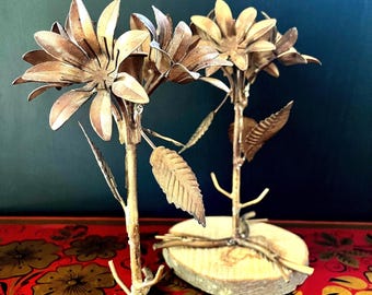 May include: Two gold metal flower sculptures with wooden bases. The flowers have delicate petals and leaves. The sculptures are on a red and black patterned surface.