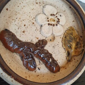May include: A small, round, ceramic dish with a brown rim. The dish has a white background with a brown branch, a white flower, and a brown and yellow leaf.