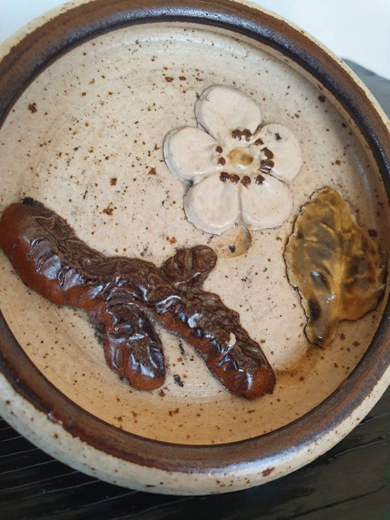 May include: A small, round, ceramic dish with a brown rim. The dish has a white background with a brown branch, a white flower, and a brown and yellow leaf.