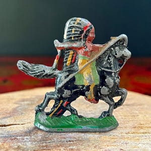 May include: A vintage toy soldier figurine of a Native American riding a black horse. The soldier is wearing a colourful headdress and holding a spear. The figurine is made of metal and painted in black, red, yellow, green, and white.