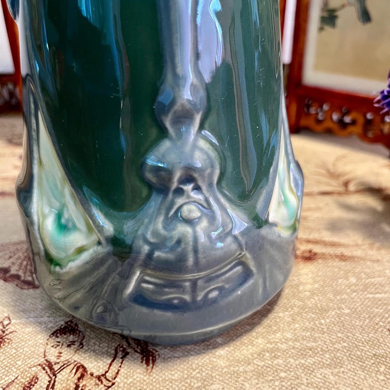 Pode incluir: Um close-up de um vaso de cer&acirc;mica com um esmalte verde escuro. O vaso tem um design em relevo detalhado em tons de azul e cinza perto da base. O design parece ser um rosto ou figura estilizada. O vaso tem um acabamento brilhante.