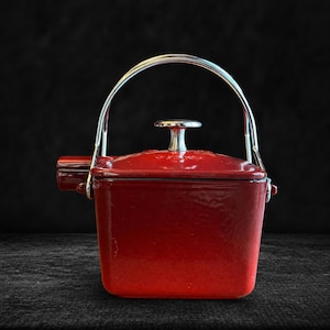 Könnte beinhalten: Ein roter emaillierter gusseiserner Bräter mit Edelstahlgriff und Deckel. Der Topf hat eine quadratische Form mit abgerundeten Ecken und einem kleinen Ausguss. Der Griff wölbt sich über die Oberseite.
