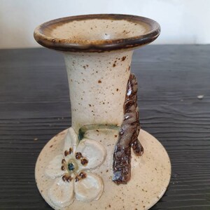May include: A ceramic candle holder with a white flower and brown branch design. The holder is beige with brown speckles.