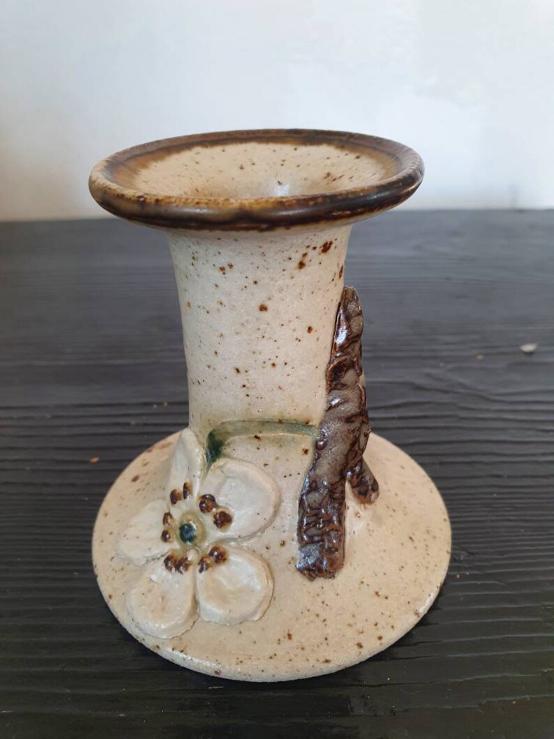May include: A ceramic candle holder with a white flower and brown branch design. The holder is beige with brown speckles.