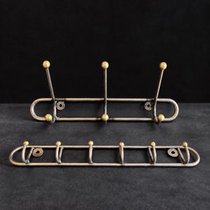 Könnte beinhalten: Zwei Wandgarderoben aus dunkelgrauem Metallrohr mit messingfarbenen, kugelförmigen Endstücken an jedem Haken. Die obere Garderobe hat drei Haken, die untere sechs Haken, beide verfügen über kreisförmige Befestigungsplatten zur Wandmontage.