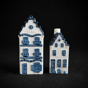 May include: Two white ceramic miniature house figurines with blue painted details. The taller house features the year 1780, while the smaller house displays the year 1627. Both structures have detailed windows, doors, and stepped gables in blue.