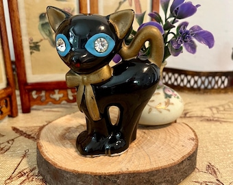 May include: A black ceramic cat figurine with gold accents, including a bow tie and tail. The cat has blue eyes with crystal centers and is standing on a wooden slice. A vintage-style decorative item.