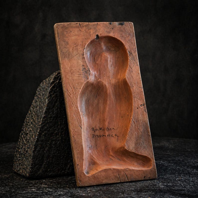 K&ouml;nnte beinhalten: Eine rechteckige, rotbraune Holzform mit einer geschnitzten menschlichen Figur. Die Form wird von einem dunklen, strukturierten Stein gest&uuml;tzt. Der Text "Sp. Nielsen Denmark" ist auf der Form eingraviert. Die Form wird wahrscheinlich zur Herstellung von Skulpturen oder Dekorationsgegenst&auml;nden verwendet.