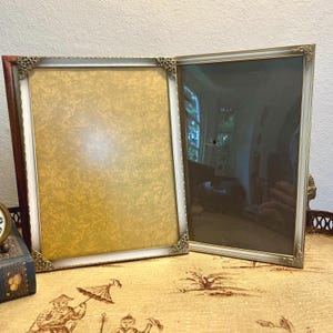 May include: Two rectangular picture frames. One frame has a brown wooden border with a white inner border and gold corner accents. The other frame has a white border with gold accents. Both frames are empty.