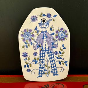 Może przedstawiać: Biała ceramiczna tablica dekoracyjna z fantazyjną ilustracją. Projekt przedstawia postać w kapeluszu i garniturze, trzymającą kompozycje kwiatowe. Postać jest w kolorze niebieskim i fioletowym, z kwiatowymi i liściastymi akcentami. U góry znajduje się otwór.