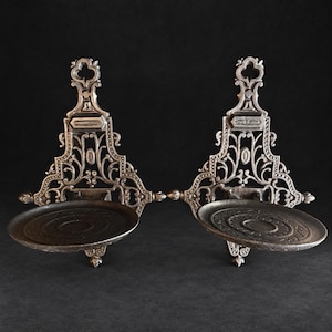 Pode incluir: Um par de castiçais de parede a condizer feitos de metal escuro. Cada peça apresenta uma placa traseira intrincada e aberta com volutas e uma base circular plana com um padrão decorativo em relevo na superfície superior.