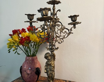 May include: A silver-toned five-arm candelabra with a cherub figure at the base. The candelabra has a decorative, ornate design and is sitting on a wooden table.