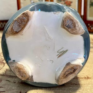 Pode incluir: Vista inferior de um vaso de cer&acirc;mica com borda verde-azulado e interior branco. O vaso tem quatro p&eacute;s castanhos e marca&ccedil;&otilde;es, incluindo os n&uacute;meros "323" e uma assinatura. O vaso &eacute; provavelmente feito &agrave; m&atilde;o.