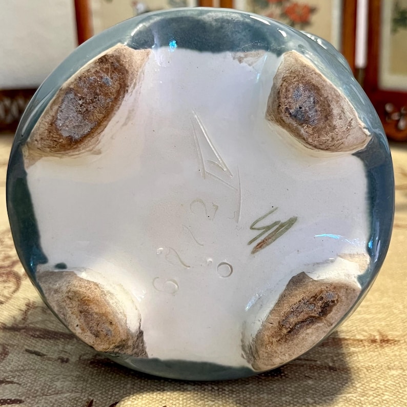 Pode incluir: Vista inferior de um vaso de cer&acirc;mica com borda verde-azulado e interior branco. O vaso tem quatro p&eacute;s castanhos e marca&ccedil;&otilde;es, incluindo os n&uacute;meros "323" e uma assinatura. O vaso &eacute; provavelmente feito &agrave; m&atilde;o.