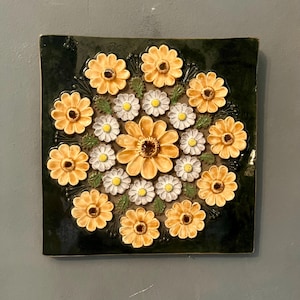 Könnte beinhalten: Quadratische Keramikfliese mit floralem Design. Die Fliese hat einen dunkelgrünen Hintergrund mit einer kreisförmigen Anordnung von gelben und weißen gänseblümchenartigen Blumen. Die Blumen haben detaillierte Blütenblätter und Zentren, wodurch ein dreidimensionaler Effekt entsteht.