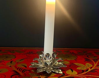 May include: A white candle with a flame burning in a silver candle holder shaped like a flower. The candle holder is sitting on a red and gold patterned surface.