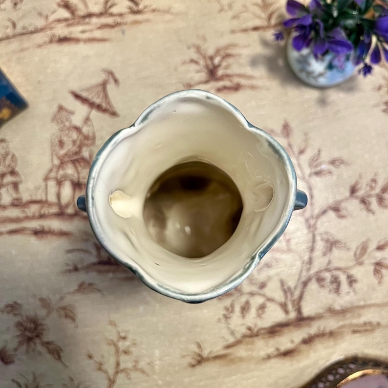 Pode incluir: Um pequeno vaso de cer&acirc;mica esbranqui&ccedil;ada com uma borda ondulada e duas pequenas al&ccedil;as. A borda &eacute; delineada em azul claro. O interior do vaso &eacute; castanho. O vaso est&aacute; sobre uma superf&iacute;cie estampada.