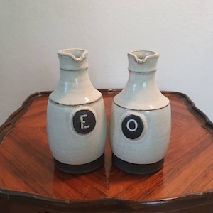 May include: Two white ceramic oil and vinegar bottles with black lids and black circles with the letters "E" and "O" on the front. The bottles are sitting on a wooden table.