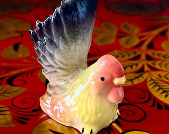 May include: A small ceramic figurine of a rooster with a yellow body, pink head, and black tail feathers. The rooster is standing on a red and gold surface with floral designs.