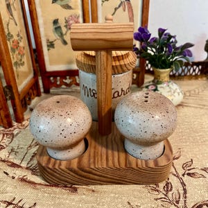 May include: A wooden salt and pepper shaker set with a matching mallet. The salt and pepper shakers are speckled, with a light brown base and a darker brown top. The set is displayed on a wooden base.