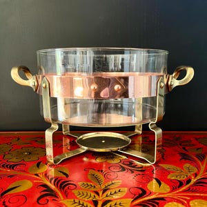 Könnte beinhalten: Eine antike Chafing Dish aus Kupfer und Glas mit Messinggriffen und einem Ständer. Die Schale hat eine klare Glasschale mit einem Kupferband um die Oberseite. Der Ständer hat eine runde Basis und vier Beine.