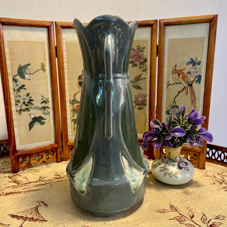 Pode incluir: Um jarro alto de cer&acirc;mica verde com uma al&ccedil;a decorativa. O jarro tem uma borda alargada e detalhes ornamentados. O fundo inclui um biombo com obras de arte florais e de p&aacute;ssaros e um pequeno vaso com flores roxas.