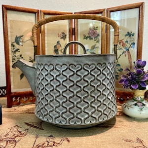 Puede incluir: Una tetera de cerámica gris con un diseño de patrón de corazón. Cuenta con un pico curvo, un pequeño asa en la tapa y un asa de ratán tejido. La tetera se exhibe frente a una pantalla plegable decorativa.