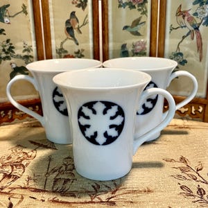May include: Three white ceramic mugs with a dark blue snowflake design. Each mug has a curved handle and a circular emblem. The mugs are arranged on a patterned surface, with a decorative screen in the background.
