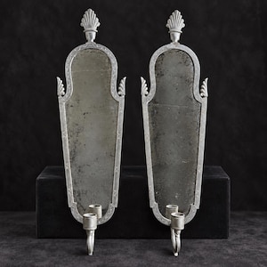 May include: A pair of silver-toned wall sconces featuring elongated, arched mirror panels with weathered, distressed surfaces. Each sconce is topped with a decorative finial and includes a single candle holder at the base with a curved support.