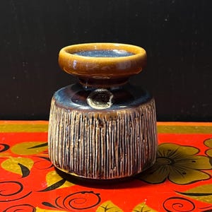 Peut inclure: Petit vase en céramique avec glaçure marron et bleue. Le dessus est de forme arrondie avec un bord marron, et le corps présente des lignes texturées verticales. Le vase repose sur une surface rouge avec des motifs floraux.