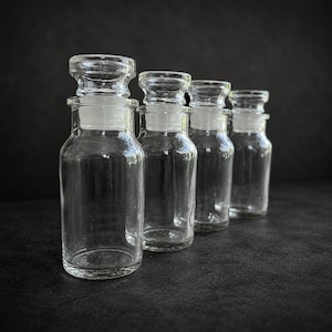 Peut inclure: Quatre bocaux d'apothicaire en verre transparent avec des bouchons en verre sont disposés en rangée. Les bocaux sont vides et de forme cylindrique, avec une base plus large et un col plus étroit. Les bouchons sont en verre transparent assorti.
