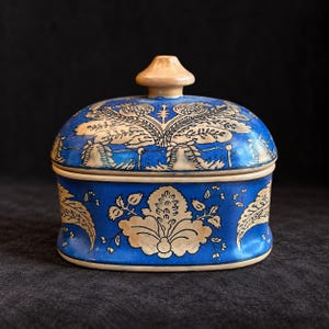 Può includere: Una scatola ovale in ceramica con coperchio, caratterizzata da una vivace smaltatura blu con intricati motivi floreali e di foglie color crema. Il coperchio è sormontato da un piccolo pomello arrotondato beige chiaro e la base ha un bordo crema.