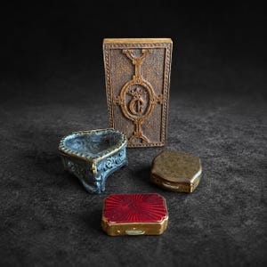 Könnte beinhalten: Eine Sammlung von vier kleinen Behältern, darunter eine hohe rechteckige Schachtel mit einem Kronen- und Buchstaben-C-Emblem, ein herzförmiges Gefäß mit verziertem Relief, eine kleine Metalldose und eine Schachtel mit rotem Strahlenmuster.