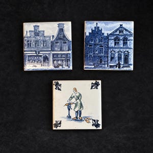 Może przedstawiać: Trzy kwadratowe płytki ceramiczne z ręcznie malowanymi wzorami. Dwie płytki przedstawiają niebiesko-białe sceny architektoniczne z wielopiętrowymi budynkami. Trzecia płytka przedstawia osobę pracującą przy narzędziu.