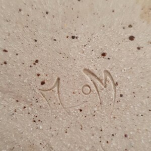 May include: Close-up of a white ceramic plate with brown speckles. The plate has a hand-painted signature that reads "MOM" in cursive.