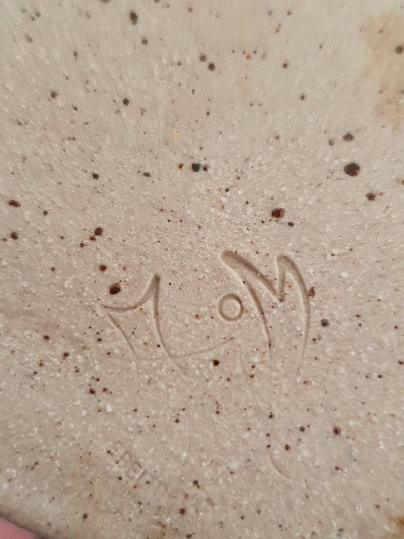 May include: Close-up of a white ceramic plate with brown speckles. The plate has a hand-painted signature that reads "MOM" in cursive.