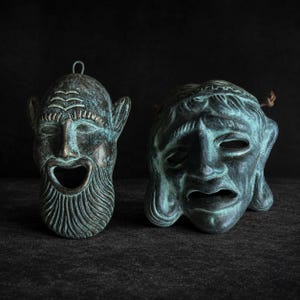 Könnte beinhalten: Zwei dekorative Wandmasken mit einer Patina aus verwitterter Bronze und Grünspan. Die linke Maske zeigt ein bärtiges Gesicht mit offenem Mund und Aufhängeöse. Die rechte Maske stellt ein trauriges Gesicht mit welligem Haar und braunen Kordeln dar.