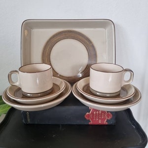 Könnte beinhalten: Zwei Sets aus braunen und weißen Keramik-Teetassen und Untertassen mit einem braunen Tablett. Die Tassen und Untertassen haben einen braunen Streifen am Rand. Das Tablett hat ein braunes Kreisdesign in der Mitte.