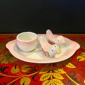 May include: A ceramic salt and pepper set featuring two bird-shaped shakers and a small cup, all resting on a pink and white oval tray. The birds are light pink with yellow beaks and green accents.