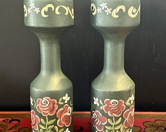 May include: A pair of green wooden candle holders with hand-painted floral designs. Each holder features red roses, white flowers, and green leaves. The holders have a yellow and white decorative band around the top.