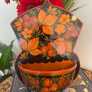 Op de afbeelding: Een zwart en oranje geschilderde houten wandzak met een komvormige bodem. De zak heeft een bloemmotief met rode bessen en oranje bladeren. De kom is geschilderd met een vergelijkbaar bloemmotief.