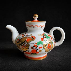 May include: A white porcelain teapot featuring a hand-painted design of a rooster and floral motifs in vibrant orange, green, and metallic gold. The teapot has a curved handle, a short spout, and a lid topped with a small, gold-accented knob.
