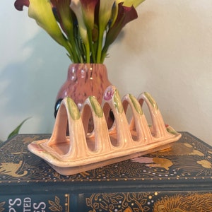 May include: A vintage pink ceramic toast rack with six arched slots, decorated with green and pink floral details. It rests on a decorative book, with a vase of calla lilies in the background.
