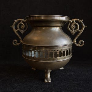 Puede incluir: Una urna de metal bronceado con asas ornamentadas y una banda decorativa de arcos. La urna tiene un cuerpo redondeado y se apoya en tres pies decorativos. Las asas presentan diseños florales y de volutas.