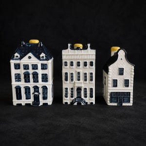Pode incluir: Três recipientes de cerâmica em forma de casa com paredes brancas, telhados azul-escuros e detalhes de janelas. Cada estrutura apresenta uma pequena tampa amarela no topo. Os designs representam diferentes estilos arquitetónicos com janelas variadas.